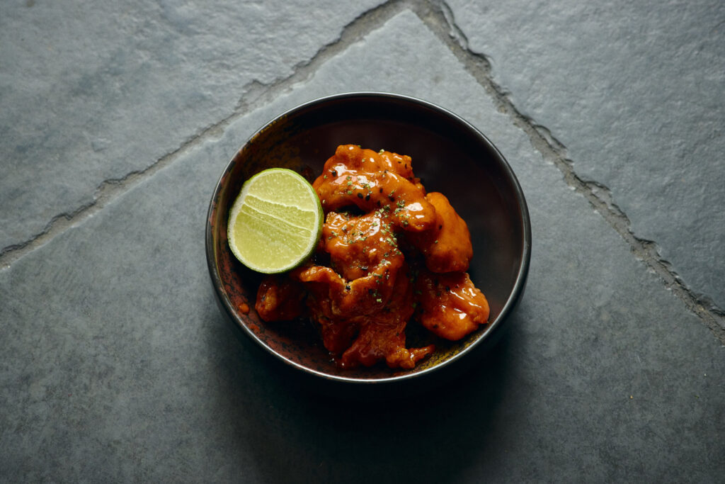 Gilpin Spice's spice fried chicken coated in a rich chilli sauce, served with fresh lime in a dark ceramic bowl