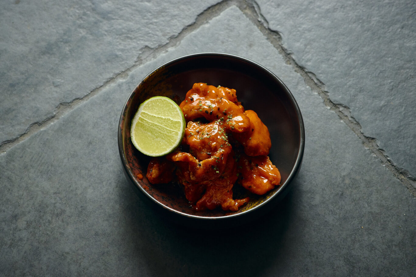 Gilpin Spice's spice fried chicken coated in a rich chilli sauce, served with fresh lime in a dark ceramic bowl