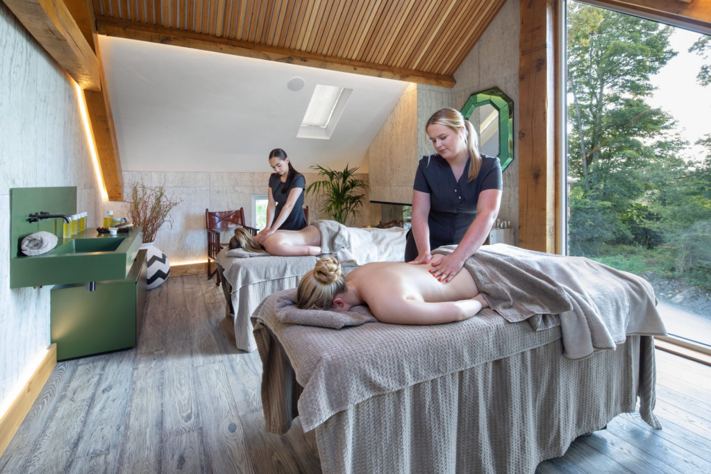 Two spa therapists giving massages in a bright treatment room at Gilpin, with large windows overlooking the surrounding woodland.