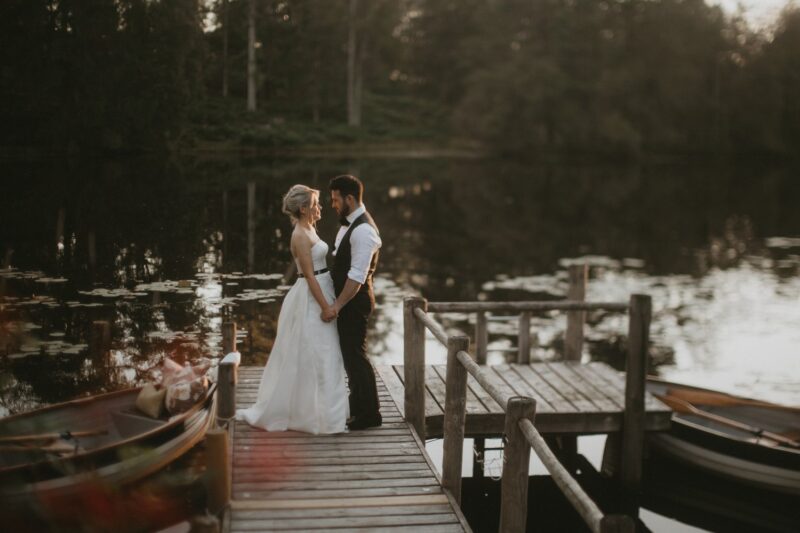 Bride and groom embracing on a wooden jetty beside the lake at Gilpin Lake House, surrounded by woodland and rowing boats.