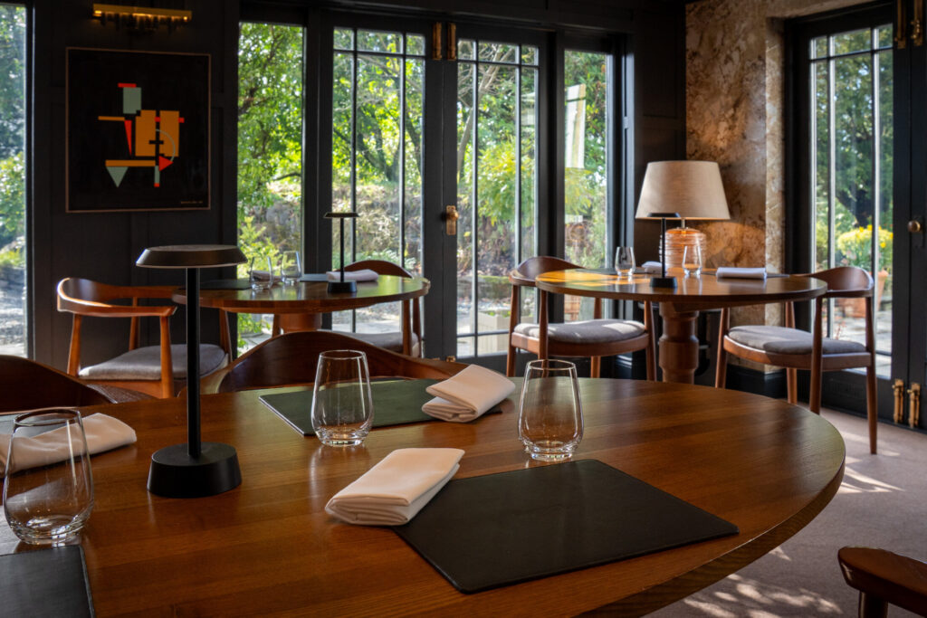 SOURCE dining room at Gilpin Hotel in summer, with tables set beside large windows overlooking the green gardens.