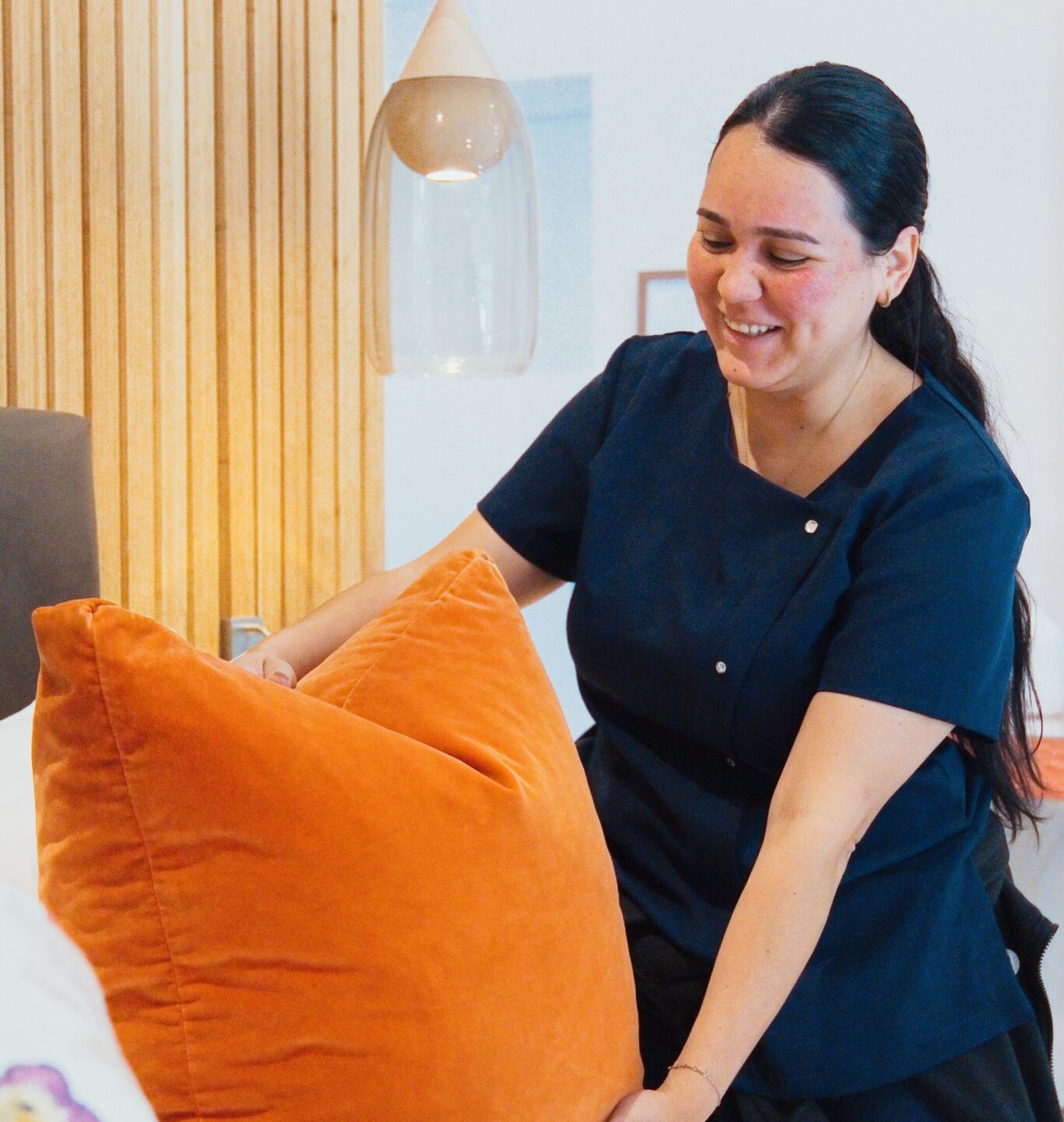 Gilpin Hotel housekeeper smiling as she arranges orange cushions on a freshly made bed in a guest room.