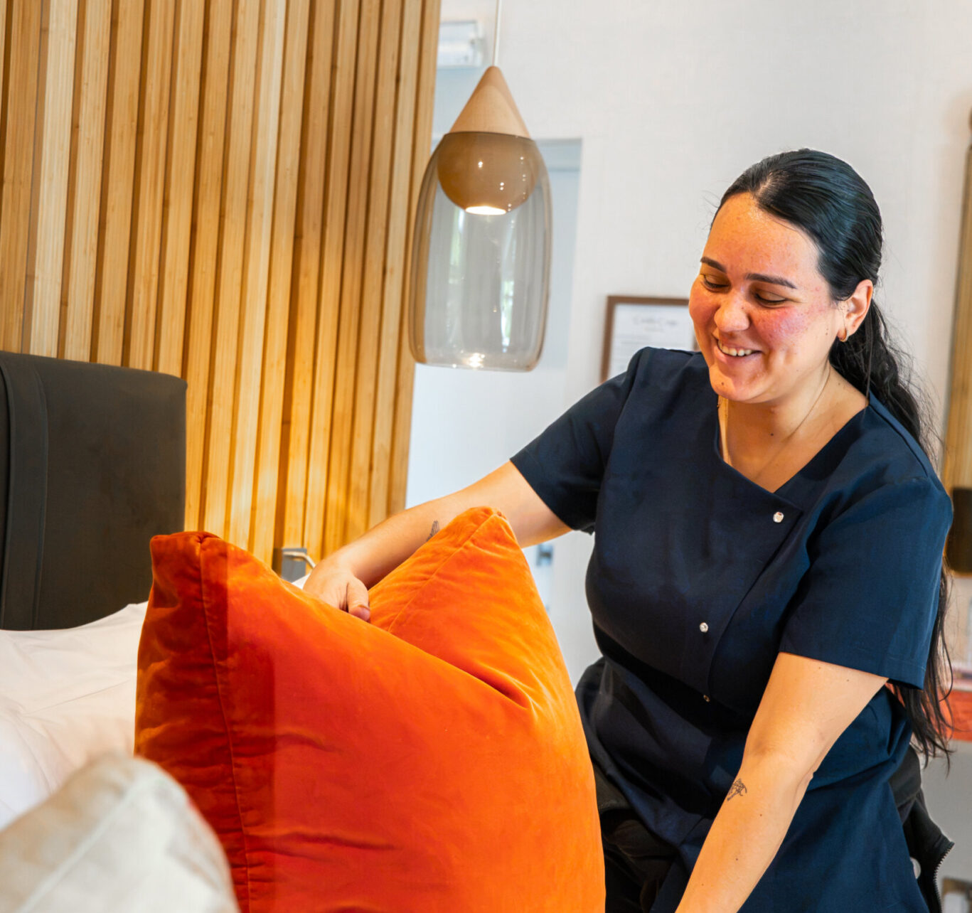 Gilpin Hotel housekeeper smiling as she arranges orange cushions on a freshly made bed in a guest room.