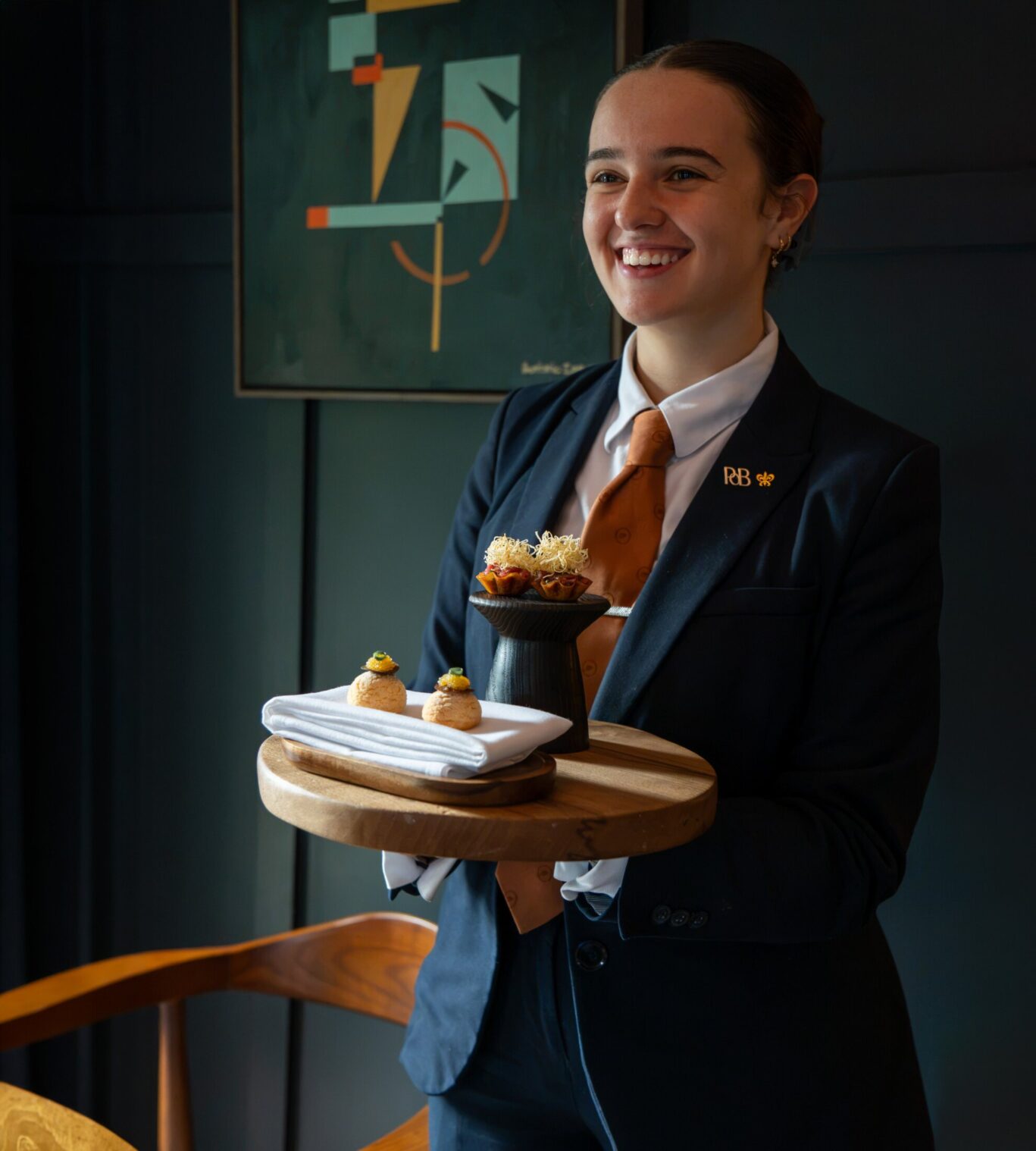 Smiling woman from the SOURCE front of house team holding a wooden tray of canapés in the restaurant at Gilpin Hotel.