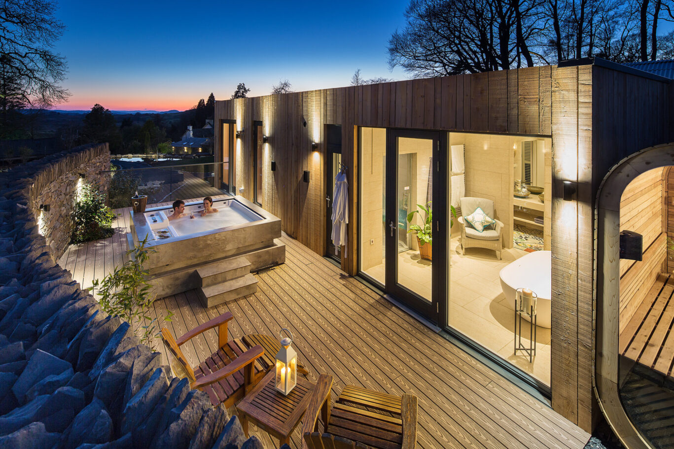 Couple relaxing in a private hot tub on the terrace of a Spa Lodge at dusk, with an outdoor sauna and illuminated bathroom.