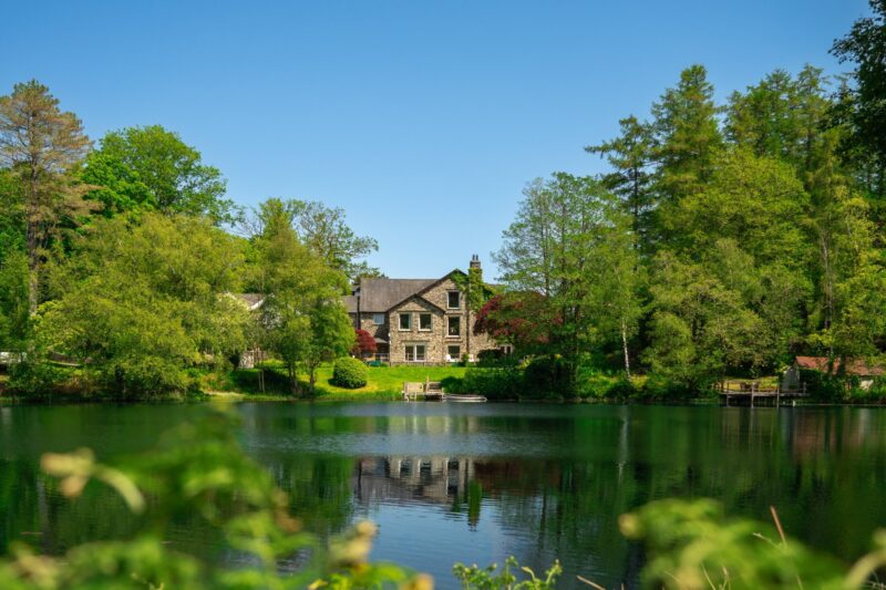 Gilpin Lake House is surrounded by woodland, viewed across the peaceful lake on a bright summer day.