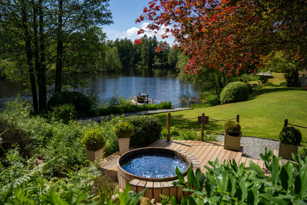 Wooden hot tub overlooking Knipe Tarn at Gilpin Lake House, surrounded by green gardens, woodland and a red-leaved tree.