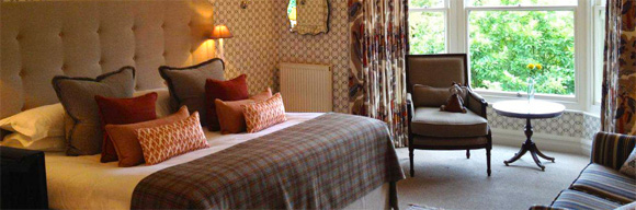Lyth Master Room at Gilpin Hotel with a king-size bed, warm patterned décor and seating beside a large window overlooking the gardens.