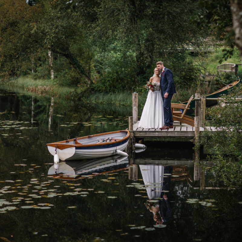 Bride and groom embracing on a private lakeside jetty at Gilpin Lake House, with a rowing boat on the water beside them.