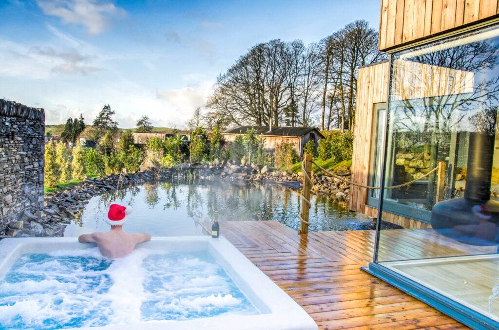 Man wearing a Santa hat relaxing in a private Spa Lodge hot tub overlooking a natural pond and woodland at Gilpin Hotel