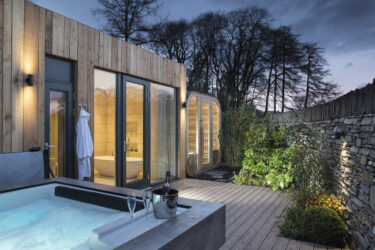 Spa Lodge exterior with a private hot tub, outdoor sauna and garden terrace, with a freestanding bath visible through the glass doors.