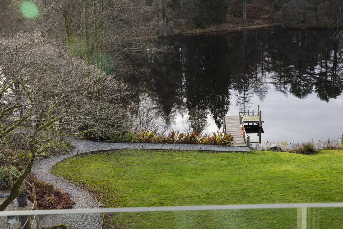 View across the gardens at Gilpin Lake House to the private lake and wooden jetty, surrounded by peaceful woodland.